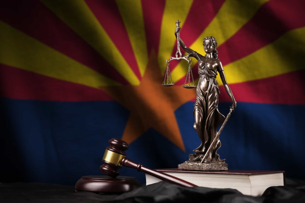 Lady Justice statue with scales, judge’s gavel, and law book in front of Arizona state flag